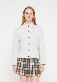 Jasnoszary dziergany kardigan z wysokim kołnierzem, z dużymi ciemnymi guzikami z przodu i ściągaczami na mankietach, zestawiony z kratowaną spódnicą.