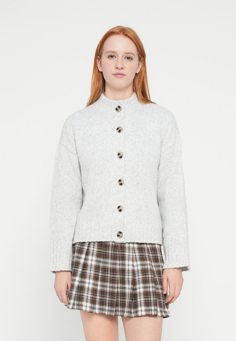 Jasnoszary dziergany kardigan z wysokim kołnierzem, z dużymi ciemnymi guzikami z przodu i ściągaczami na mankietach, zestawiony z kratowaną spódnicą.