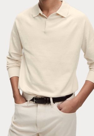 Une personne portant une chemise col montant beige à manches longues, rentrée dans un pantalon beige avec une ceinture noire, les mains dans les poches avant.