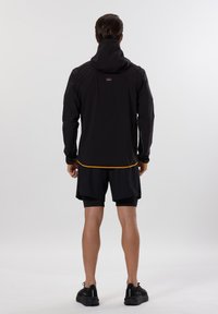 Sort vandtæt jakke med hætte, med orange detaljer ved kanten og manchetterne, båret over sorte shorts og sorte sneakers.