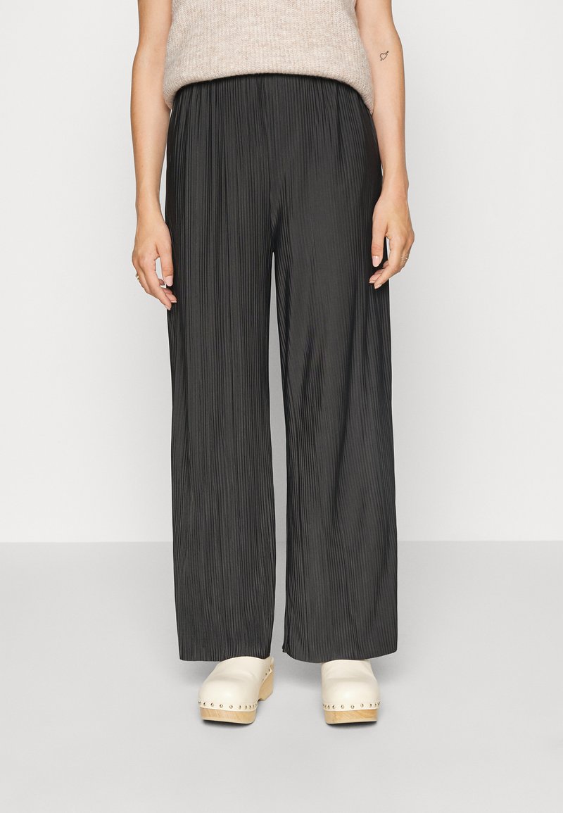 Pantaloni largi negri plisati, confecționați dintr-un material texturat, cu talie elastică, asortați cu saboți bej. Fără modele sau accente vizibile.