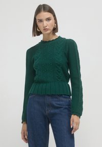 Frau mit kurzen braunen Haaren, die goldene Ohrringe, einen dunkelgrünen Strickpullover mit gewellten Kanten und blaue Jeans trägt, vor einem schlichten Hintergrund.