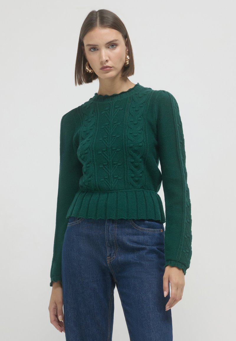 Frau mit kurzen braunen Haaren, die goldene Ohrringe, einen dunkelgrünen Strickpullover mit gewellten Kanten und blaue Jeans trägt, vor einem schlichten Hintergrund.