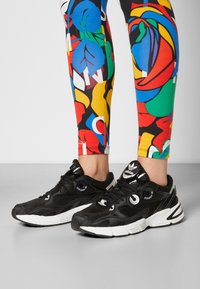 Tênis de desporto pretos com um cabedal de malha e borracha, apresentando detalhes brancos, um logo redondo e cordões texturizados, combinados com leggings coloridas e estampadas.