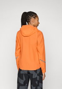 Orange wasserdichte Jacke mit Kapuze, ausgestattet mit reflektierenden Akzenten an den Ärmeln, leichtem Material und einem lässigen Schnitt.