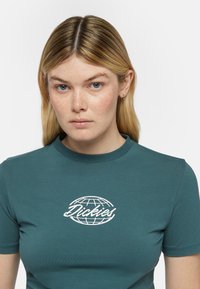 Teal lyhythihainen t-paita, joka on valmistettu sileästä kankaasta ja jossa on valkoinen maapallon logo, jossa on teksti "Dickies" keskitettynä rinnassa.