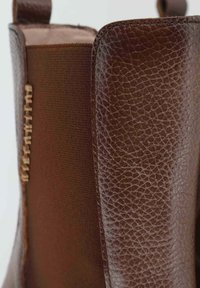 Stivale in pelle marrone con finitura testurizzata, pannello laterale elastico e un manicotto per il sollevamento. Presenta dettagli di cucitura a contrasto lungo la cucitura.