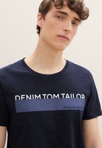 Marineblå bomulls t-skjorte med korte ermer, rund hals, med hvit tekst "DENIM TOM TAILOR" og et horisontalt strukturert stripemønster.