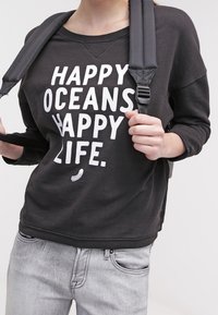 Felpa nera con testo bianco "OCEANI FELICI VITA FELICE", scollatura ampia, design corto e tessuto a coste. Jeans grigi indossati sotto.