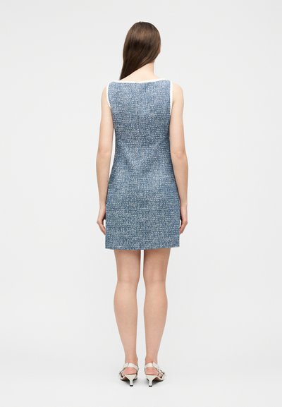 Femme debout de dos, portant une robe en tweed bleu sans manches et des sandales blanches à talons, sur un fond uni.