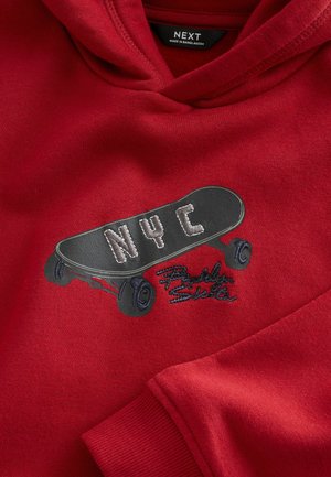 Punainen huppari, jossa on musta skeittilautagrafiikka ja valkoisilla ja sinisillä yksityiskohdilla varustettu "NYC". Pinta näyttää pehmeältä; kangas on todennäköisesti puuvillasekoitetta.