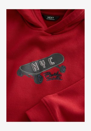 Sweat à capuche rouge arborant un graphique de skateboard noir avec "NYC" en blanc et des détails bleus. La texture semble douce ; le tissu est probablement un mélange de coton.