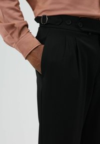 Zwarte op maat gemaakte broek met plooien, voorzien van een riem met een zilveren gesp en knoopsluiting. Gecombineerd met een lichtroze blouse.