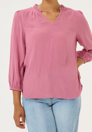 Woman wearing a pink long-sleeve blouse with a ruffled V-neck collar and light blue jeans against a plain background.