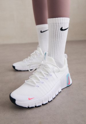 Hvide Nike sneakers med lyserøde og blå accenter, båret med hvide Nike sokker på et beton gulv.