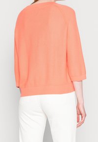 Maglione in maglia corallo dal taglio morbido, con maniche a tre quarti, orlo a costine e un motivo testurizzato, indossato con pantaloni bianchi.