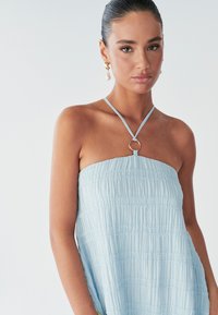 Model draagt een strapless, lichtblauwe jurk met een geplooide textuur en een ronde ringdetail bij de halslijn, met natuurlijke make-up en oorbellen.
