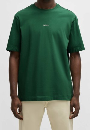 Homme portant un t-shirt vert foncé à manches courtes avec le mot « BOSS » imprimé en petits caractères blancs sur la poitrine et un pantalon beige.