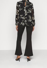 Femme portant un chemisier à manches longues noir à motif floral, un pantalon évasé noir et des mules à talons hauts noires, debout de dos sur un fond uni.