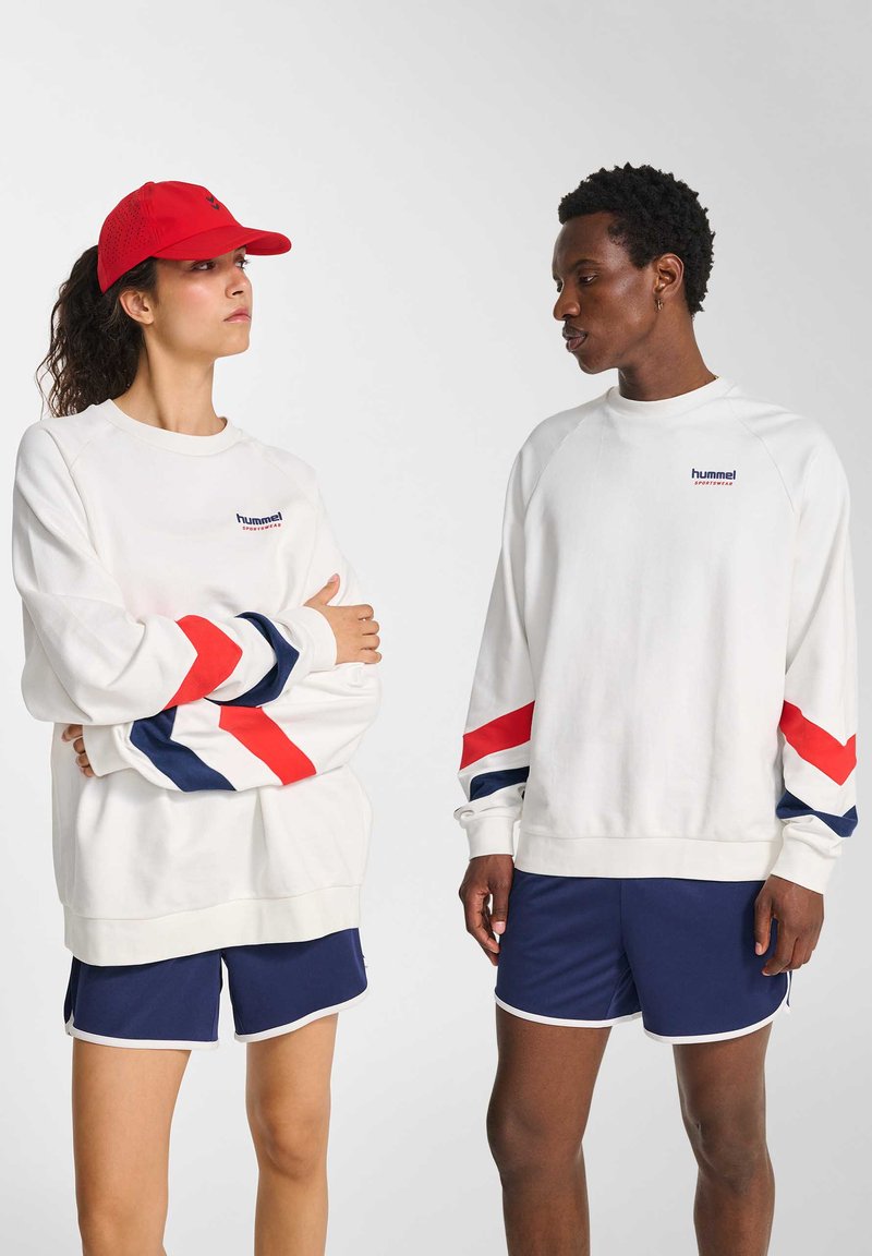 To personer iført hvide sweatshirts med røde og blå chevronærmer og marineblå shorts, vendt mod hinanden mod en ensfarvet baggrund.