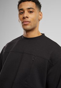 Czarny sweatshirt z wysokim kołnierzem, teksturowanym materiałem i małą kieszonką na zamek błyskawiczny na klatce piersiowej. Charakteryzuje się luźnym krojem i minimalistycznym designem.