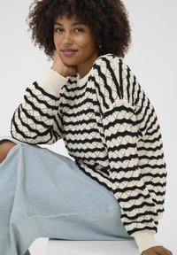 Un pull en tricot noir et crème présentant un motif ondulé et des poignets côtelés. Le modèle porte un pantalon en denim bleu clair, mettant en avant une coupe décontractée.