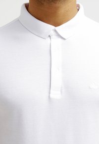 Vit polo skjorta med ett texturerat tyg, en tre knapps placket och en vikt krage. Har en subtil logo på vänster sida.
