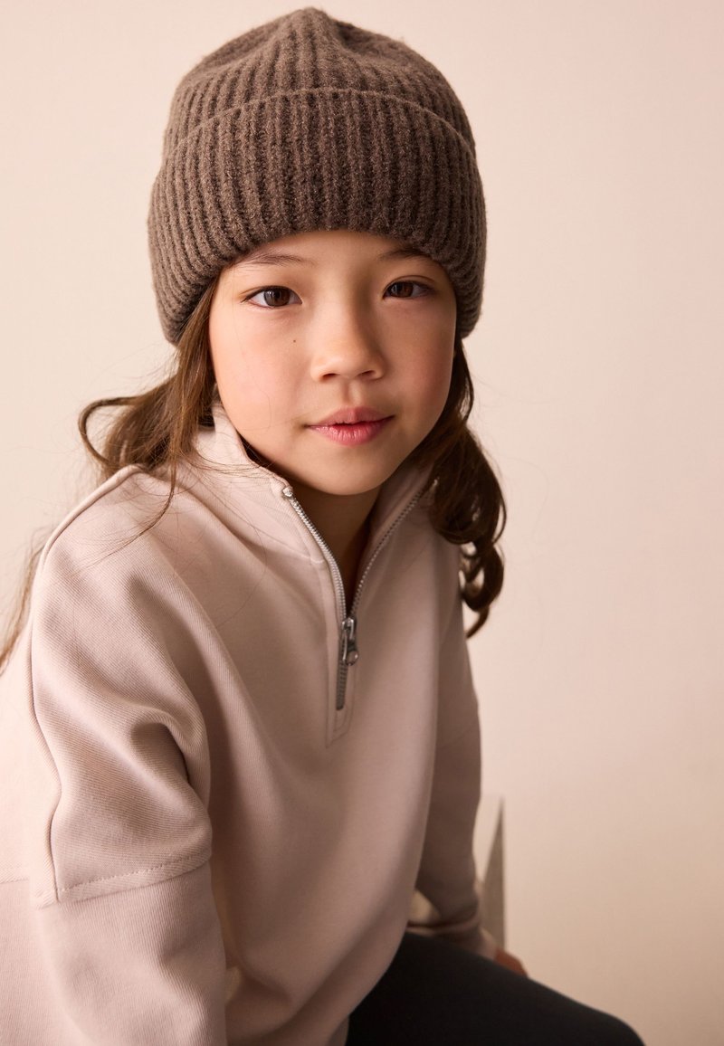 Child wearing a brown ribbed knit beanie and a light pink zip-up sweatshirt. Background is a soft neutral color. Hair is wavy and dark.