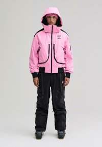 Ung person iført en lyserød hættejakke, matchende lyserød hue, sorte cargobukser og snestøvler, stående foran en ensfarvet baggrund.