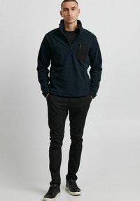Schwarzer Reißverschluss-Pullover mit hohem Kragen, aus strukturierter Stoff und mit einer Brusttasche. Kombiniert mit schwarzen Slim-Fit-Hosen und schwarzen Sneakers.