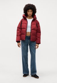 Chaqueta acolchada roja con capucha, cremallera negra y cordones, llevada encima de un top corto blanco y unos jeans de pierna ancha azules. Unos zapatos negros completan el look.