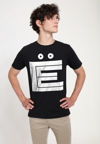 Černé tričko s velkým bílým grafickým designem připomínajícím písmeno "E" se dvěma kulatýma očima nahoře. Bavlněný materiál.