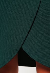 Tyrkysová zavinovací sukně vyrobená z texturované tkaniny, s diagonálním lemem a vrstveným designem pro přidanou hloubku.