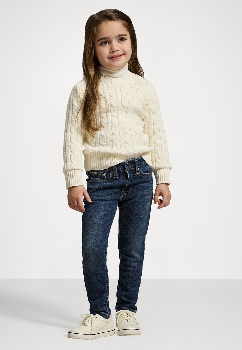 Suéter de punto cableado color blanco roto, jeans oscuros y zapatillas blancas. Niña se presenta con confianza, con cabello largo castaño y piel clara.