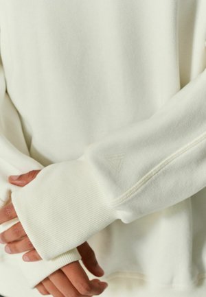 Personne portant un pull blanc à manches longues avec des trous pour les pouces, les mains doucement croisées à la taille, montrant la texture douce en tricot et les détails des coutures.