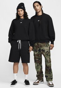 Μαύρα φούτερ και cargo σορτς σε μαύρο και καμουφλάζ; αξεσουάρ περιλαμβάνουν μαύρο σκουφάκι και αθλητικά παπούτσια με λευκές και καφέ λεπτομέρειες.