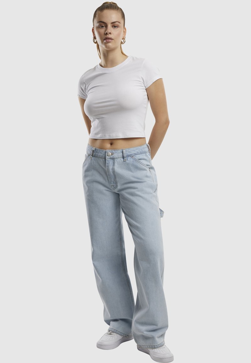 Frau steht mit den Händen in den Taschen, trägt ein eng anliegendes weißes Crop-Top, weite hellblaue Jeans, weiße Sneaker und goldene Creolen.