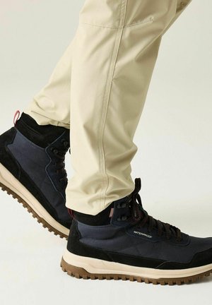 Person wearing beige pants and black waterproof hiking boots with thick brown tread soles on a light background.