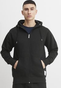 Hoodie zippé noir en tissu doux, avec une capuche, des poches avant et des cordons de serrage ajustables. Texture lisse et poignets ajustés.