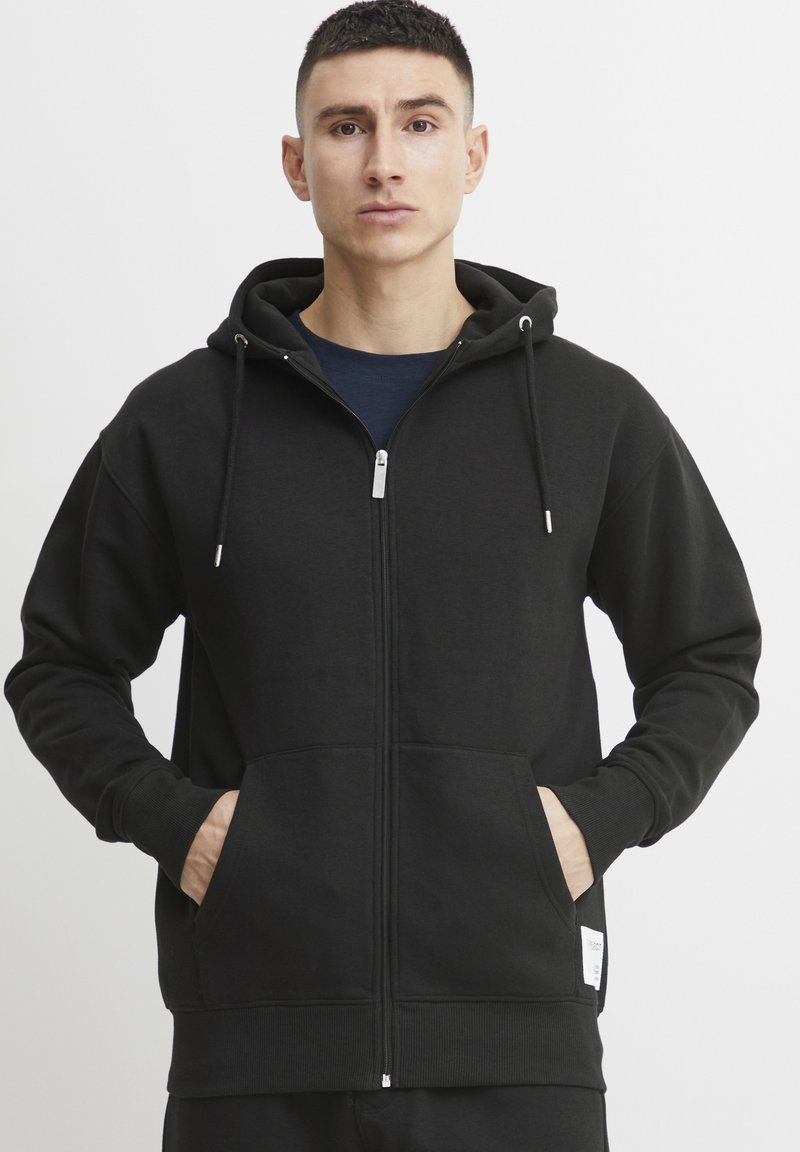 Hoodie zippé noir en tissu doux, avec une capuche, des poches avant et des cordons de serrage ajustables. Texture lisse et poignets ajustés.
