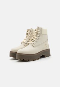 Beige leather ankle boots with round toe, lace-up front, and padded collar. Features a chunky brown rubber sole with pronounced treads.