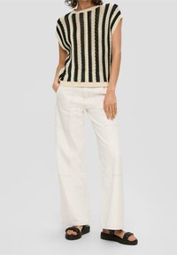 Kobieta ubrana w czarno-biały bezrękawnik w paski, białe szerokie spodnie i czarne sandały na platformie, stojąca z rękami w kieszeniach.