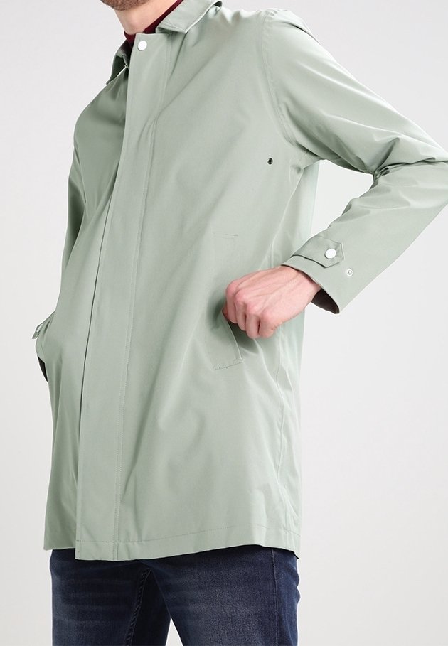 Mann trägt einen hellgrünen langen Mantel mit Druckknöpfen, die Hand in der Tasche, steht vor einem einfarbigen Hintergrund.