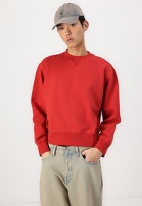Rød hettegenser med lange ermer og rund hals, med ribbestrikkede mansjetter og kant. Bærers sammen med lyseblå denimbukser og en grå caps.