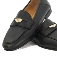 Zwarte leren loafers met een gouden decoratief embleem op de band, tan interieurvoering en lage hak, weergegeven op een witte achtergrond.