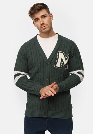 Mørkegrøn kabelstrikket cardigan med v-hals, udstyret med et hvidt "M" emblem og stribede detaljer på ærmerne, lukket med mørke knapper.