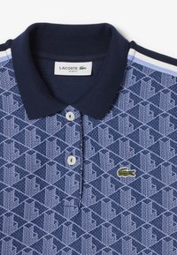 Polo shirt Lacoste ajusté à motif bleu avec col bleu foncé, boutons blancs et logo crocodile vert sur la poitrine.