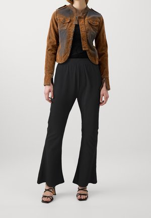 Veste courte en daim brun avec des fermetures à boutons et des poches avant, associée à un pantalon noir à jambes larges et des sandales noires à lanières.