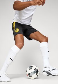 Sorte fodboldshorts med gul sidestribe og holdlogo, parret med hvide sokker og fodboldstøvler. En mønstret fodbold ligger på jorden.