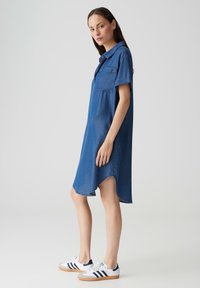 Jeanshemdkleid, kurze Ärmel, Knopfkragen, eine Brusttasche, geschwommener Saum, blaue Farbe, lockere Passform, kombiniert mit weißen Turnschuhen.